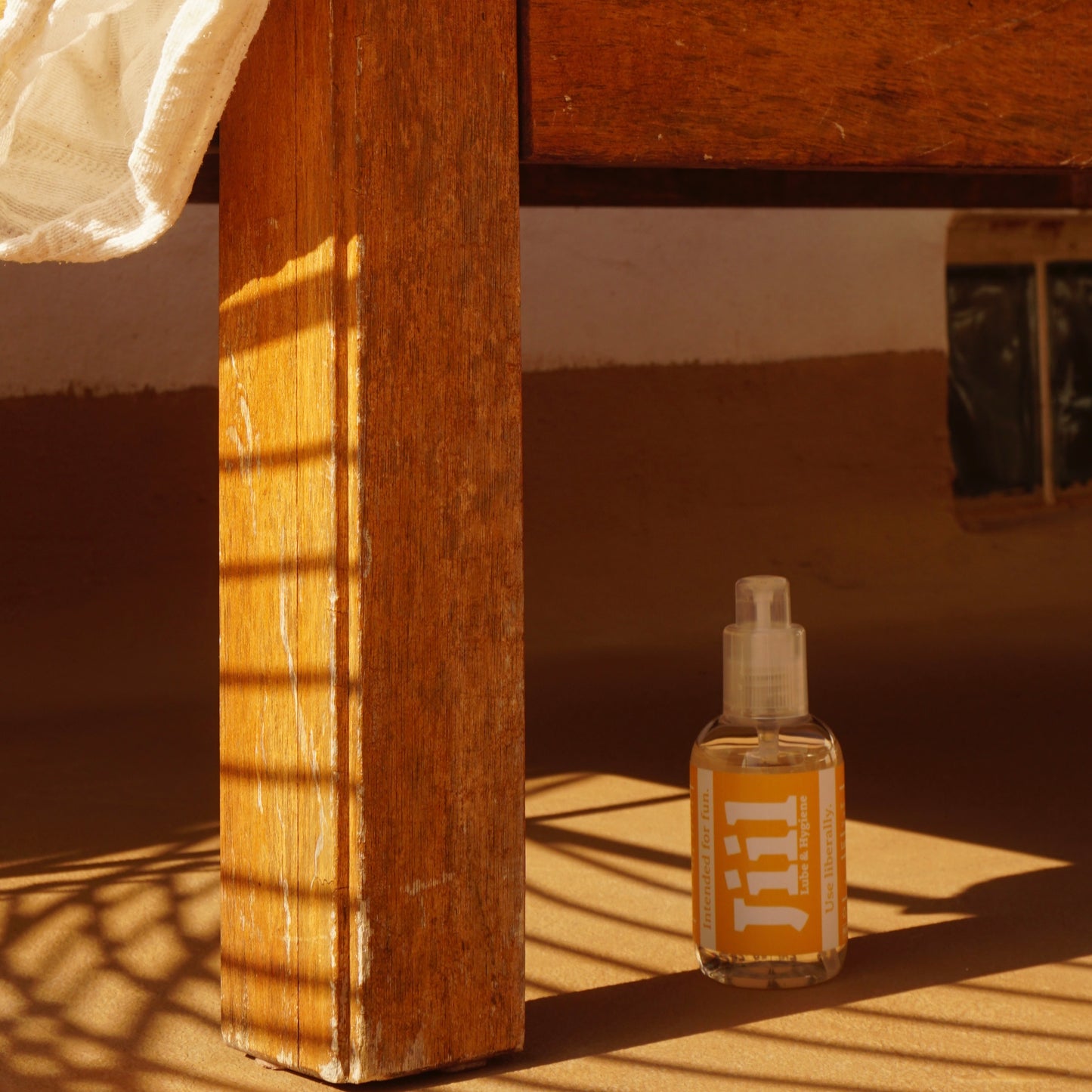 Clear plastic bottle of Jill personal lubricant with a yellow label, positioned on a shaded floor under a wooden table. Jill lube & hygiene bottle with a yellow label, sitting on a sunlit floor next to a rustic wooden table leg. Product image of Jill personal lubricant, showing the bottle under a wooden table with sunlight and shadow play.